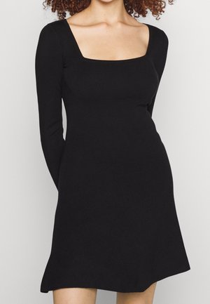 Femme portant une robe noire à manches longues, longueur genou, avec un décolleté carré et un corsage ajusté.