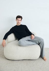 Kreslo s čalúnením v odtieni off-white, zaobleného tvaru; osoba so sladkým dlhým rukávom v čiernej farbe a sivých nohaviciach pohodlne sedí.