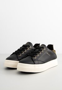 Schwarze Ledersneaker mit einer weißen Gummisohle, ausgestattet mit goldenen Ösen, schwarzen Schnürsenkeln und einem Akzent aus hellbraunem Wildleder an der Ferse. Markenlogo an der Seite.