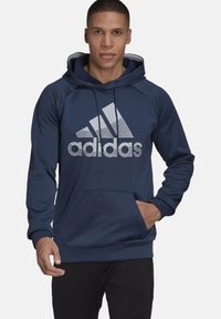 Sudadera azul marino con capucha ajustable, bolsillo canguro y gran logo gris de Adidas en la parte delantera. Tejido suave, mangas largas, corte regular.