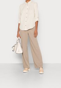 Chemise en lin beige clair à boutons avec manches retroussées, associée à un pantalon ample beige. Sac à main crème et baskets beige clair complètent le look.