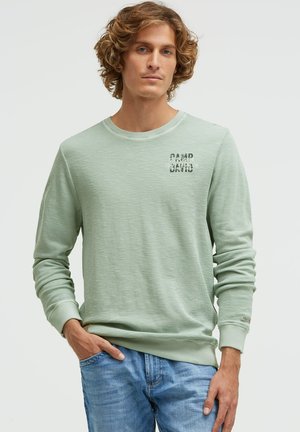 Junger Mann mit lockigem Haar, der ein hellgrünes Langarmshirt mit "Camp David"-Aufdruck und blaue Jeans trägt, Hand in der Tasche.