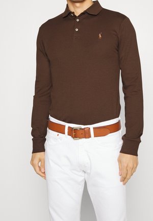 Hombre con camiseta tipo polo de manga larga marrón con logo en el pecho, pantalones blancos y cinturón de cuero color marrón claro con hebilla dorada sobre fondo blanco.