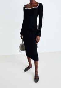 Kvinna iklädd en svart ribbad topp med lång ärm och vit halsringning, matchande asymmetrisk kjol, spikade svarta flats och håller en liten nätväska.