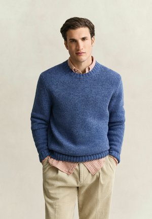 Young man with dark hair wearing a blue sweater over a red checkered shirt and beige corduroy pants, standing with hands in pockets against plain background.