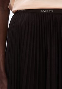 Jupe plissée noire avec une texture douce et une ceinture élastique ornée du logo "LACOSTE" en argent. Gros plan sur les détails du matériau.