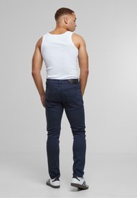 Tmavě modré slim-fit džíny s texturovaným povrchem, doplněné bílým tílkem a černým pásem. Boty mají tři bílé pruhy.