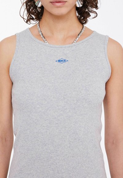Grijze geribbelde tanktop met een getailleerd ontwerp, met een blauw logo op het borstgebied en een eenvoudige ronde halslijn. Zilveren ketting als sieraad.