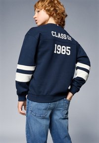 Marineblaues Sweatshirt mit "KLASSE VON 1985" in Weiß auf dem Rücken, mit weißen Streifen an den Ärmeln. Getragen mit hellblauen Jeans.