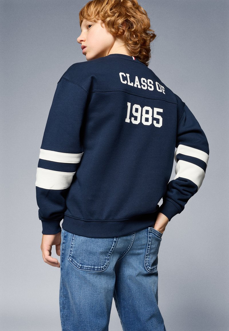 Marineblaues Sweatshirt mit "KLASSE VON 1985" in Weiß auf dem Rücken, mit weißen Streifen an den Ärmeln. Getragen mit hellblauen Jeans.