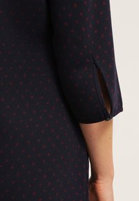 Tissu bleu foncé à petits pois rouges, présentant une demi-manche avec une fente latérale et un petit bouton noir près du poignet.