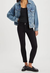 Chaqueta de mezclilla azul con un corte relajado, top negro sin mangas, jeans ajustados negros de cintura alta y mocasines negros con suelas gruesas.