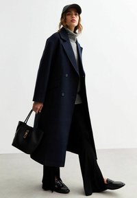 Lang marineblauwe jas met een dubbelzijdig ontwerp, gestructureerde grijze coltrui, zwarte handtas en glanzende zwarte loafers. Eenvoudige achtergrond.