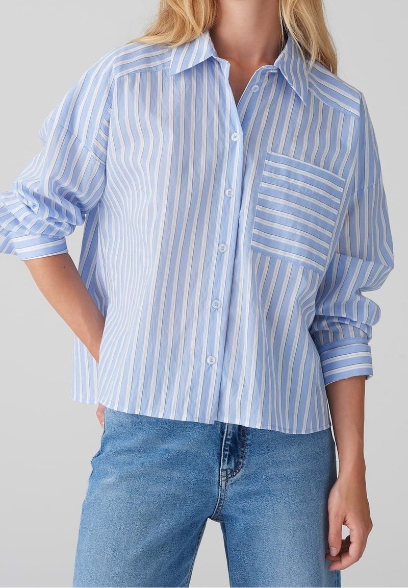 Femme portant une chemise ample à rayures verticales bleues et blanches, manches retroussées, et un jean bleu clair.