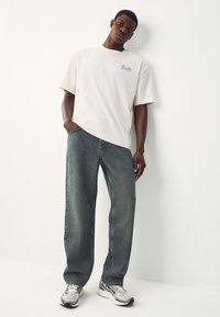 Cream short-sleeve t-shirt with purple logo, paired with loose-fit, faded blue jeans, complemented by gray sneakers with black accents.