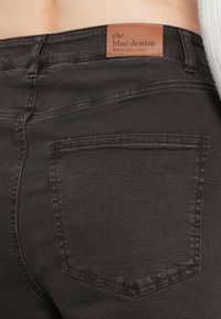 Zwarte denim jeans met een zichtbare achterzak, inclusief een leren label in bruin met de tekst "the blue denim" op de taille.