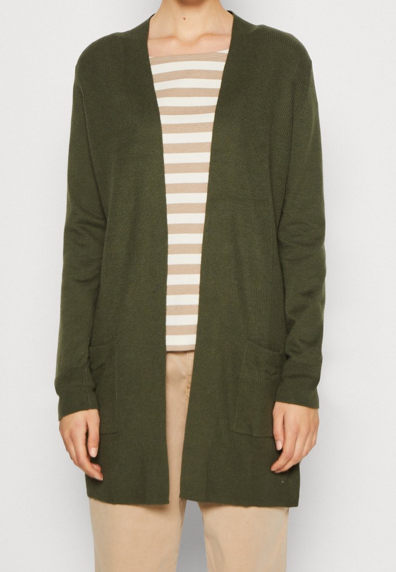 Personne portant un cardigan en maille vert foncé ouvert sur une chemise beige et blanche à rayures avec un pantalon beige, debout devant un fond uni.