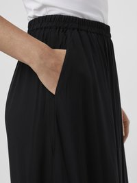Pantalon avec taille élastique noire et poche latérale, présentant une coupe ample et un tissu fluide, mis en valeur par une texture douce et un drapé lisse.
