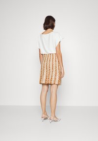 Haut blanc à manches courtes et jupe orange à motifs, présentant des rayures verticales ressemblant à un feuillage. Le mannequin porte des chaussures à talons blanches.