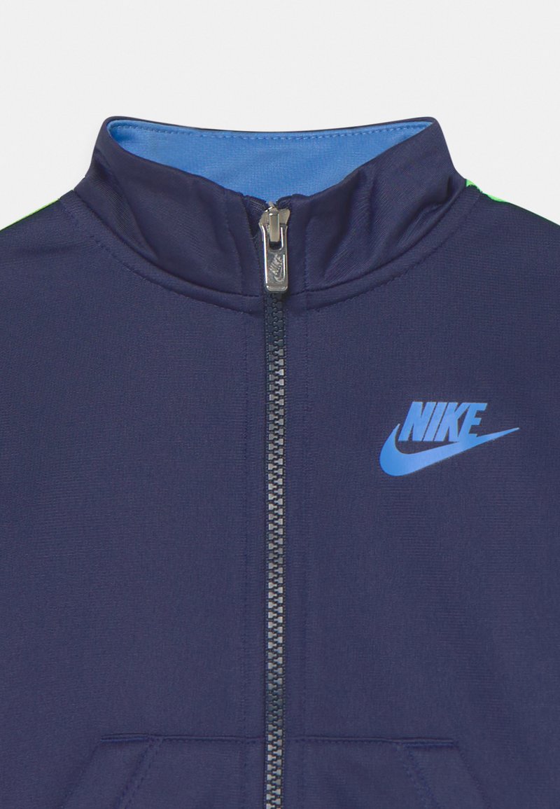 Chaqueta deportiva de color azul marino con cremallera y cuello alto, con un logo de Nike azul y un forro interior en contraste también azul. Textura de tela suave.