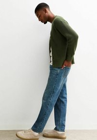 Cardigan verde a coste, camicia bianca sotto, jeans blu in denim, sneakers beige. Profilo in piedi con la mano sinistra in tasca, sfondo neutro.