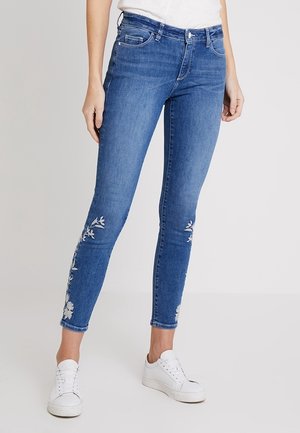 Vaqueros azul ajustados con bordado floral blanco en las piernas inferiores, combinados con zapatillas blancas y una camiseta blanca metida por dentro.
