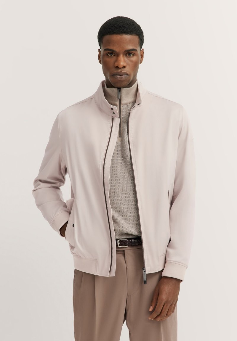 Hellrosa Jacke mit Stehkragen, Reißverschluss, seitlichen Taschen und gerippten Bündchen, kombiniert mit einem beigen Pullover und beigen Hosen.