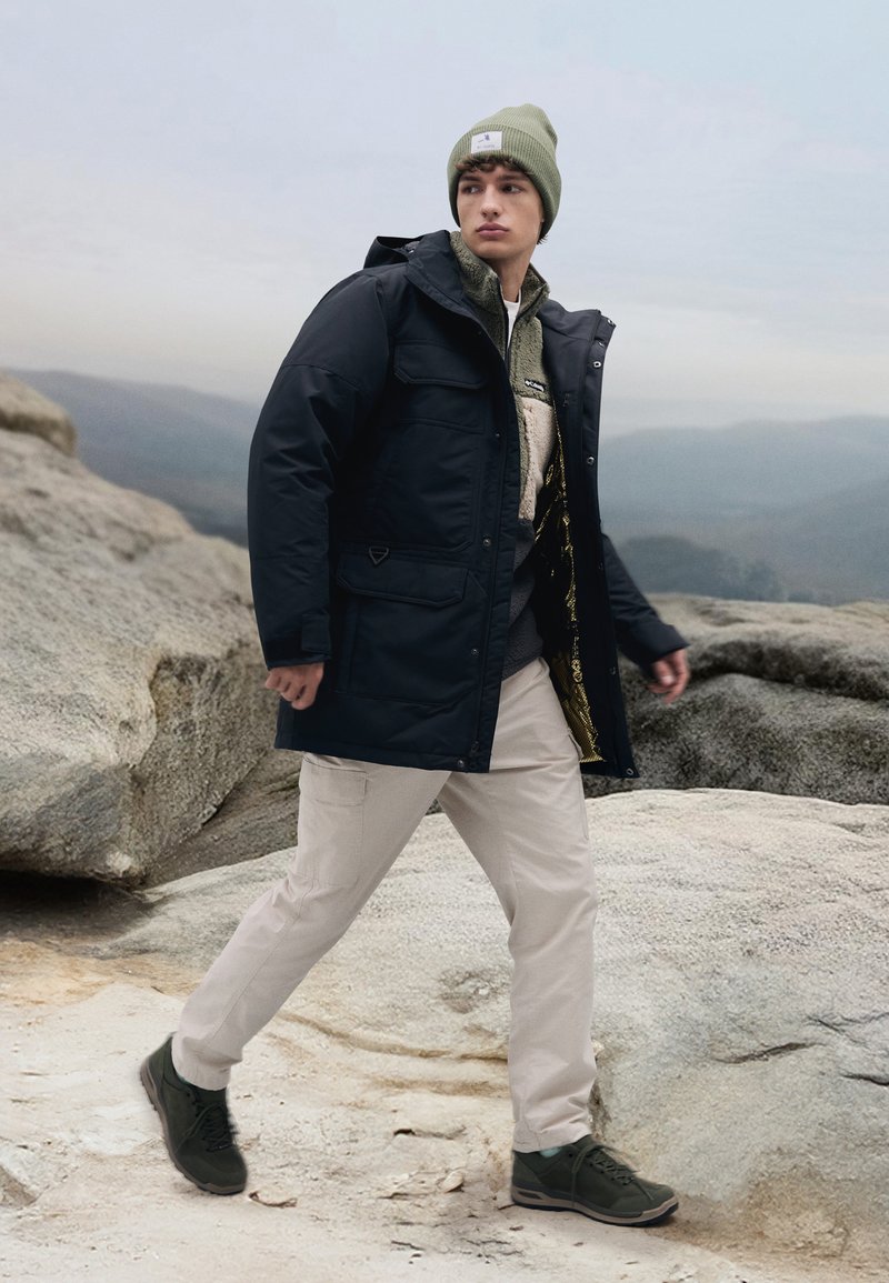 Zwarte geïsoleerde jas met meerdere zakken, gelaagd over een lichte trui, beige cargobroek en donkergroene sneakers. Model draagt een groene beanie.