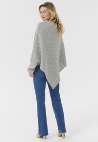 Ljust grått stickat poncho med ett texturerat sicksackmönster och asymmetrisk nederkant, buren över blå jeans och matchad med beiga klackar.