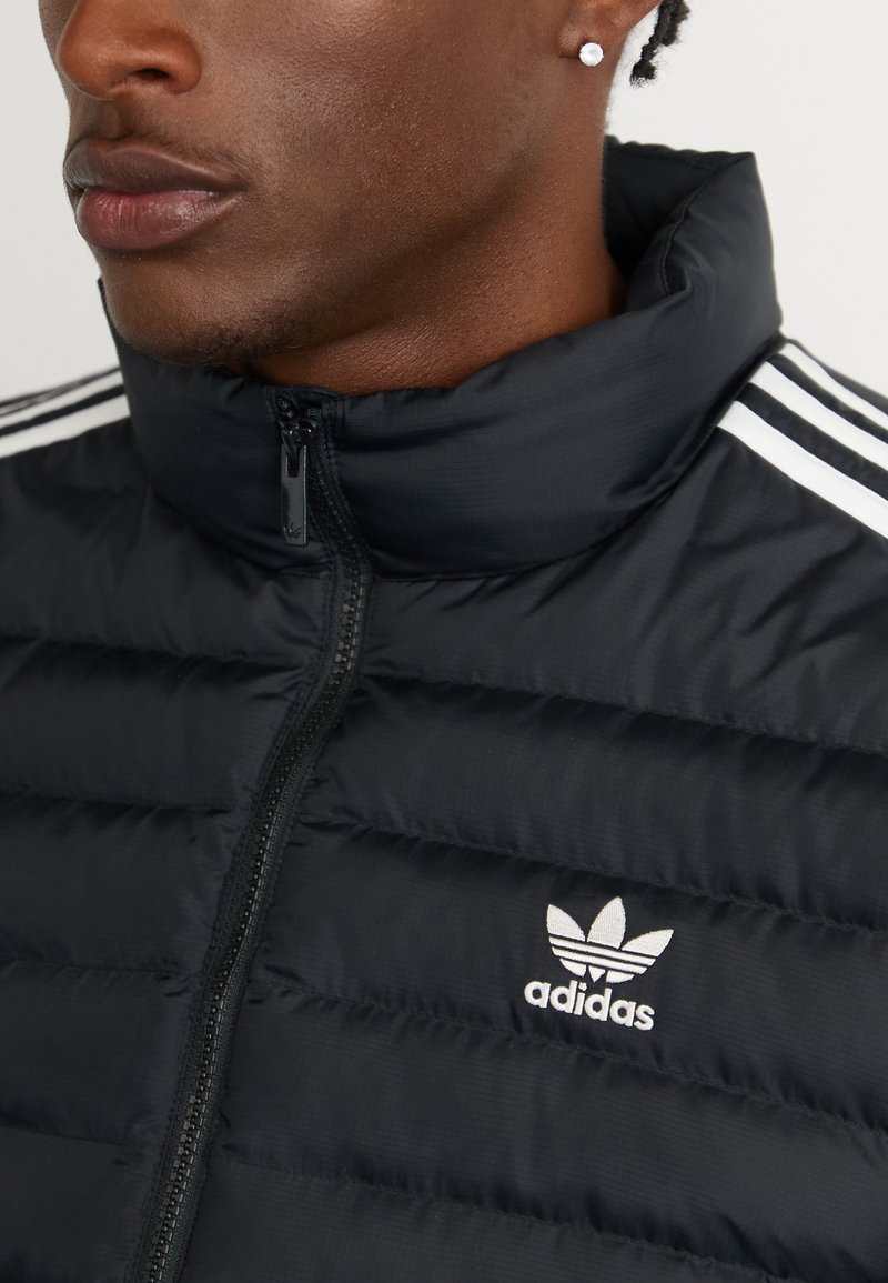 Zbliżenie osoby w czarnej kurtce puchowej Adidas z białym logo i paskami oraz małym okrągłym kolczykiem w lewym uchu.