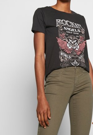 T-shirt noir à manches courtes avec un motif de crâne ailé rose et un texte, porté avec un pantalon ajusté vert olive sur une personne debout.