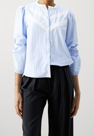 Vrouw draagt een lichtblouse met blauwe strepen en witte kanten details, gecombineerd met zwarte, hooggetailleerde, geplooide broek, tegen een effen achtergrond.