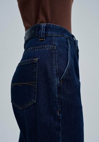 Jeans de mezclilla de un azul oscuro con una cintura alta, con costuras naranjas y un diseño de bolsillo trasero. Incluye una etiqueta de cinturón de cuero negro.