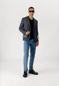 Gray blazer over a black shirt, blue jeans, and black shoes. Accessories include a gray shoulder bag and dark sunglasses. Plain background.