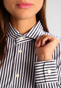 Schwarz-weiße gestreifte Bluse mit hohem Kragen, mit weißen Knöpfen und einer glatten Stofftextur. Die Manschette hat passende Streifen.