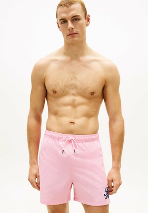 Młody muskularny mężczyzna w różowych kąpielówkach ze sznurkiem, stojący na jednolitym białym tle.
