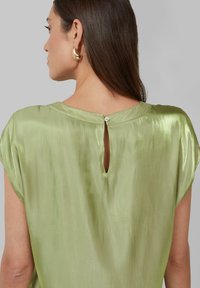 Grüne Satinbluse mit rundem Ausschnitt, kurzen Ärmeln und Knopfverschluss im Rücken mit einem Schlüsselloch-Ausschnitt.