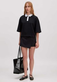 Zwarte top met korte mouwen en een witte kraag, gecombineerd met een zwarte wikkelrok. Het model houdt een glanzende zwarte tote bag vast, vergezeld van platte schoenen.