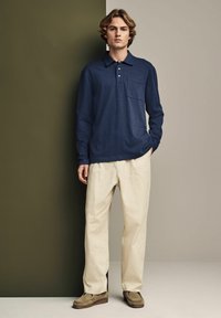 Dunkelblaues Langarm-Polohemd mit Taschen, kombiniert mit hellbeigen Hosen und olivgrünen Schuhen, vor einem neutralen Hintergrund.