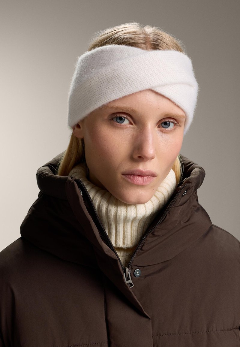 Fascia di lana bianca lavorata a maglia con un design attorcigliato, indossata con un maglione a collo alto color crema e un giubbotto imbottito marrone. Texture liscia e aspetto caldo.