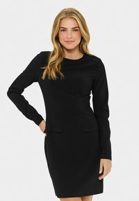 Femme ayant de longs cheveux blonds, portant une robe noire ajustée à manches longues, se tenant avec une main sur la hanche devant un fond blanc.