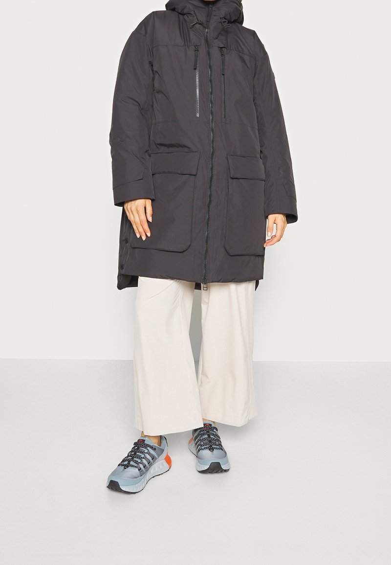 Personne portant un long parka noir à capuche, un pantalon large beige et des chaussures de sport grises avec des accents orange, debout sur un sol gris.