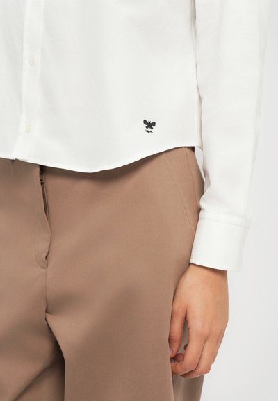 Chemise blanche à boutons avec des manches longues, dotée d'un logo de papillon noir brodé près de l'ourlet. Associée à un pantalon beige.