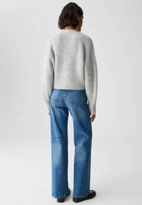Persona che indossa un maglione chunky grigio chiaro e jeans a gamba larga blu, in piedi con la schiena rivolta verso un fondo semplice e chiaro.