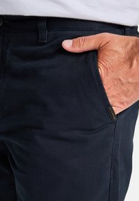 Marineblauwe katoenen broek met een gladde textuur, voorzien van zijzakken en een klein merkteken nabij de tailleband.