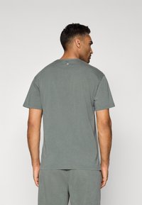 Pelēks kokvilnas T-krekls ar īsām piedurknēm, apaļu izgriezumu un smalku logotipa detalizāciju uz aizmugures. Mīksta tekstūra un standarta piegriezums.