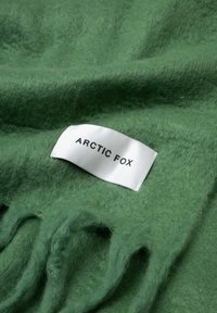 Grüner Wollteppich mit weicher Textur und franjierter Kante. Ein weißes Etikett mit schwarzem Text trägt die Aufschrift "ARCTIC FOX."