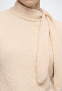 Gros plan d'un pull en maille beige douce avec un col haut et un long détail d’écharpe lâche drapé sur le côté droit.