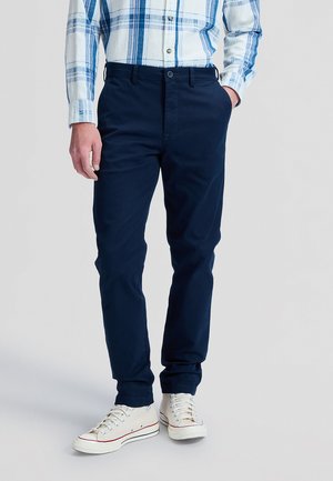 Mann mit marineblauer Hose, weißen Sneakers und einem blau-weiß karierten Hemd, steht mit einer Hand in der Tasche.