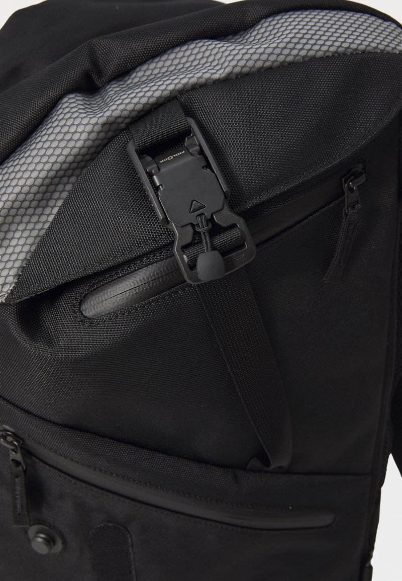 Sac à dos noir en tissu texturé, doté d'une large fermeture à boucle et de plusieurs poches zippées, y compris une poche latérale supplémentaire.